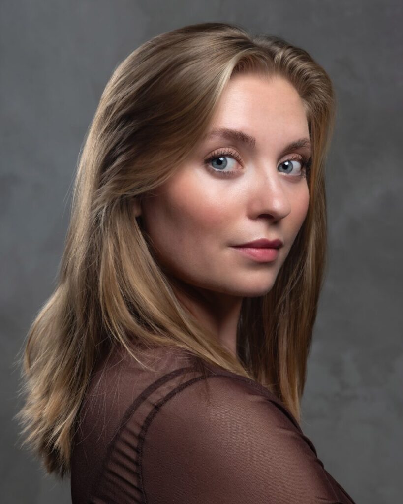 A woman photographed from the side, turning her head back over her shoulder to face the camera directly. She has long, straight, light brown hair and pale gray-blue eyes, against a plain mottled gray studio backdrop. She's wearing a sheer dark brown mesh top. The lighting is soft and directional, coming from one side.